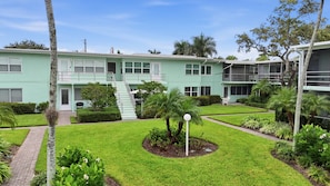 A two-story building with a light green exterior, a central courtyard with a palm tree, and a small garden area. - Downtown Naples Walkable Escape – Private Lanai, Steps to Beach & 5th Avenue (Naples)