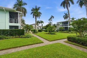 A well-maintained courtyard with a central pathway, surrounded by lush greenery and palm trees, leading to a building with a modern design. - Downtown Naples Walkable Escape – Private Lanai, Steps to Beach & 5th Avenue (Naples)