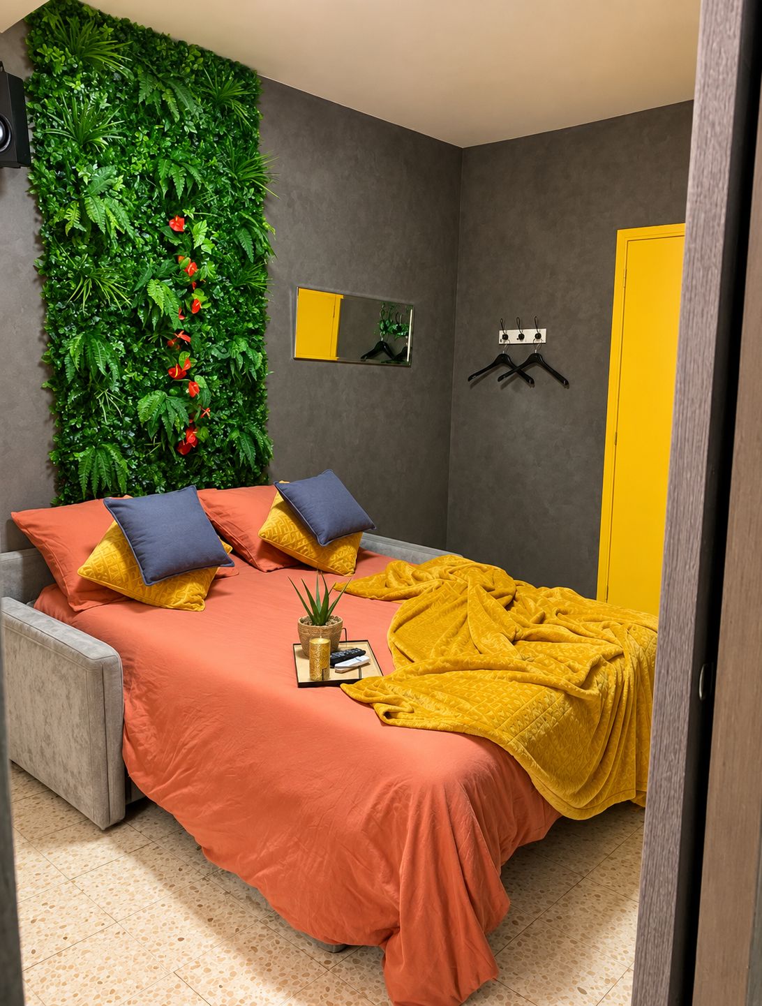 Un dormitorio con una cama, almohadas, una colcha, un espejo en la pared y una pared verde con plantas.