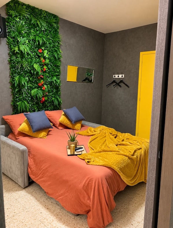 Een slaapkamer met een bed, kussens, een deken, een vastgeplakt spiegel en een groene muur met planten.