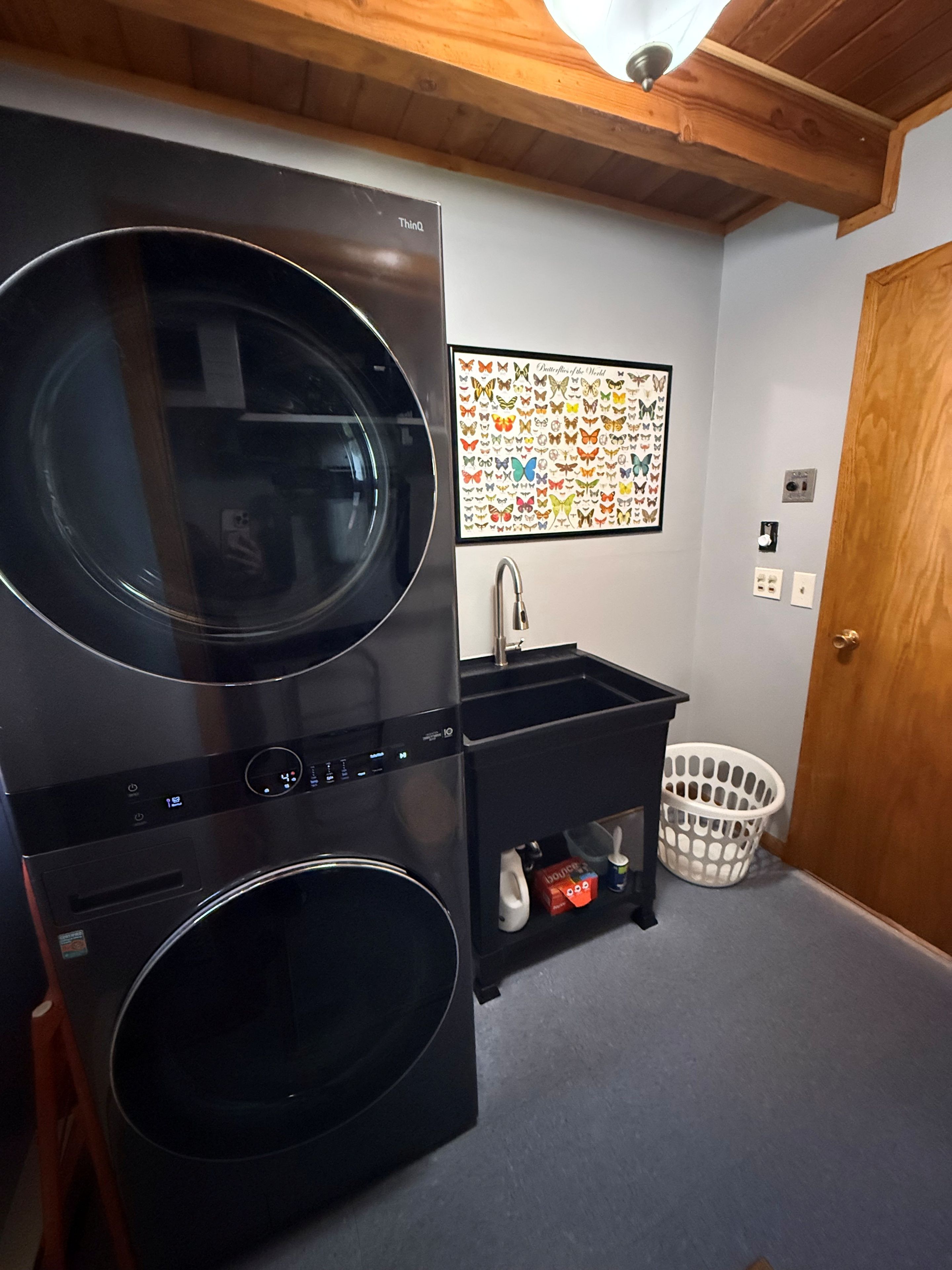 A laundry room with a stacked washer and dryer, a small black utility cabinet, a wall-mounted picture of butterflies, and a wooden door.