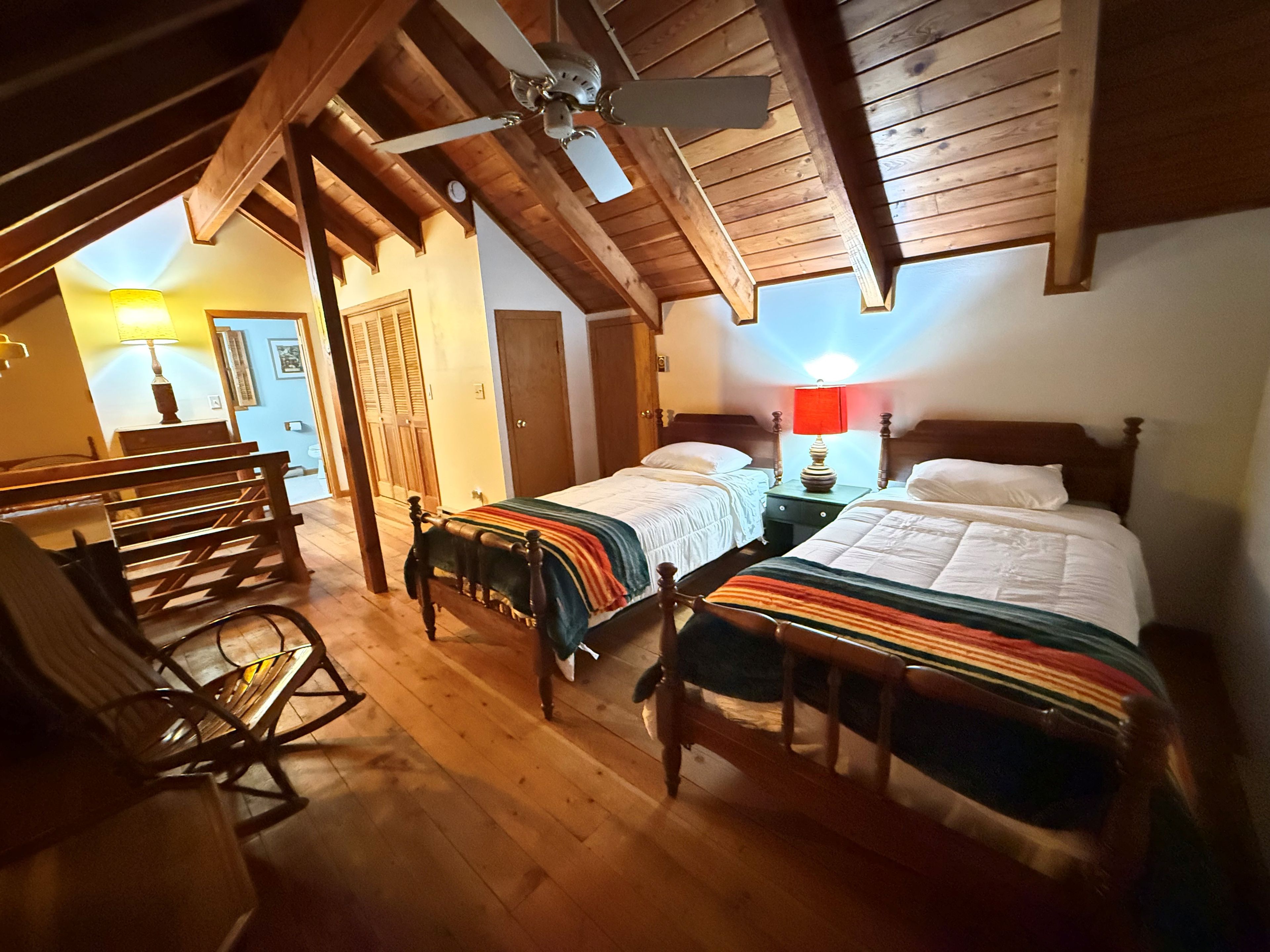 A room with two beds, wooden flooring, a rocking chair, and a ceiling fan.