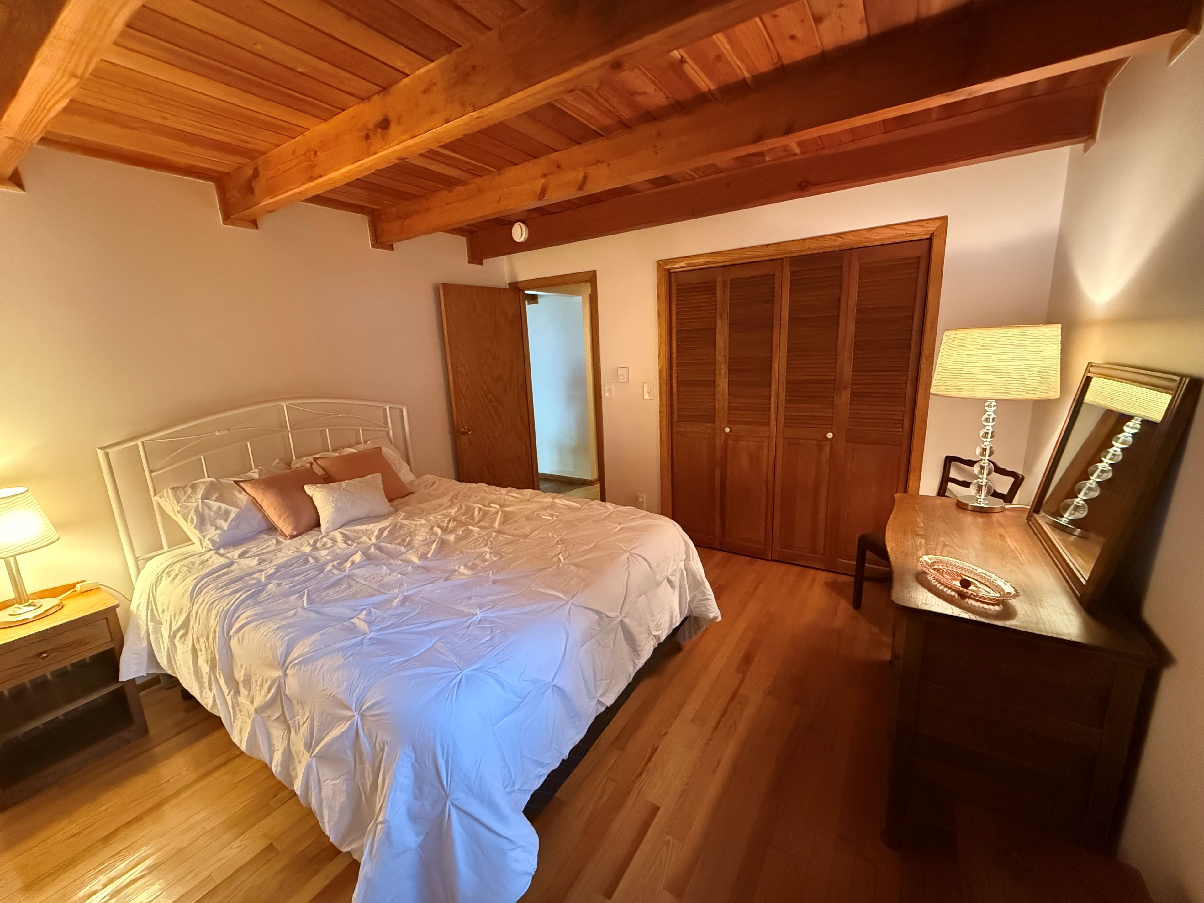 A bedroom with a wooden ceiling, a bed with white bedding, a bedside table with a lamp, and a wooden wardrobe.