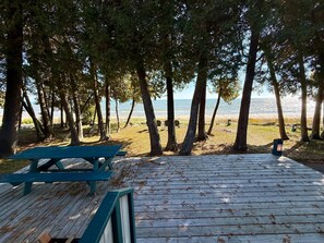 A wooden deck with a picnic table and benches overlooking a beach with trees and a clear view of the water. - Jacksonport Sandy Beach Cottage (Baileys Harbor)
