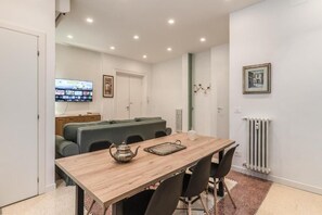 A modern living room with a wooden dining table, black chairs, a green sofa, a television mounted on the wall, and a radiator. - Buccari Apartment - 3 Bedrooms with En-suite Bathrooms - Near the Vatican (Roma)