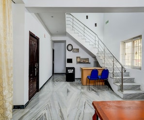 Un vestíbulo de hotel con piso de mármol, mostrador de recepción de madera, sillas azules, una escalera y un reloj en la pared.