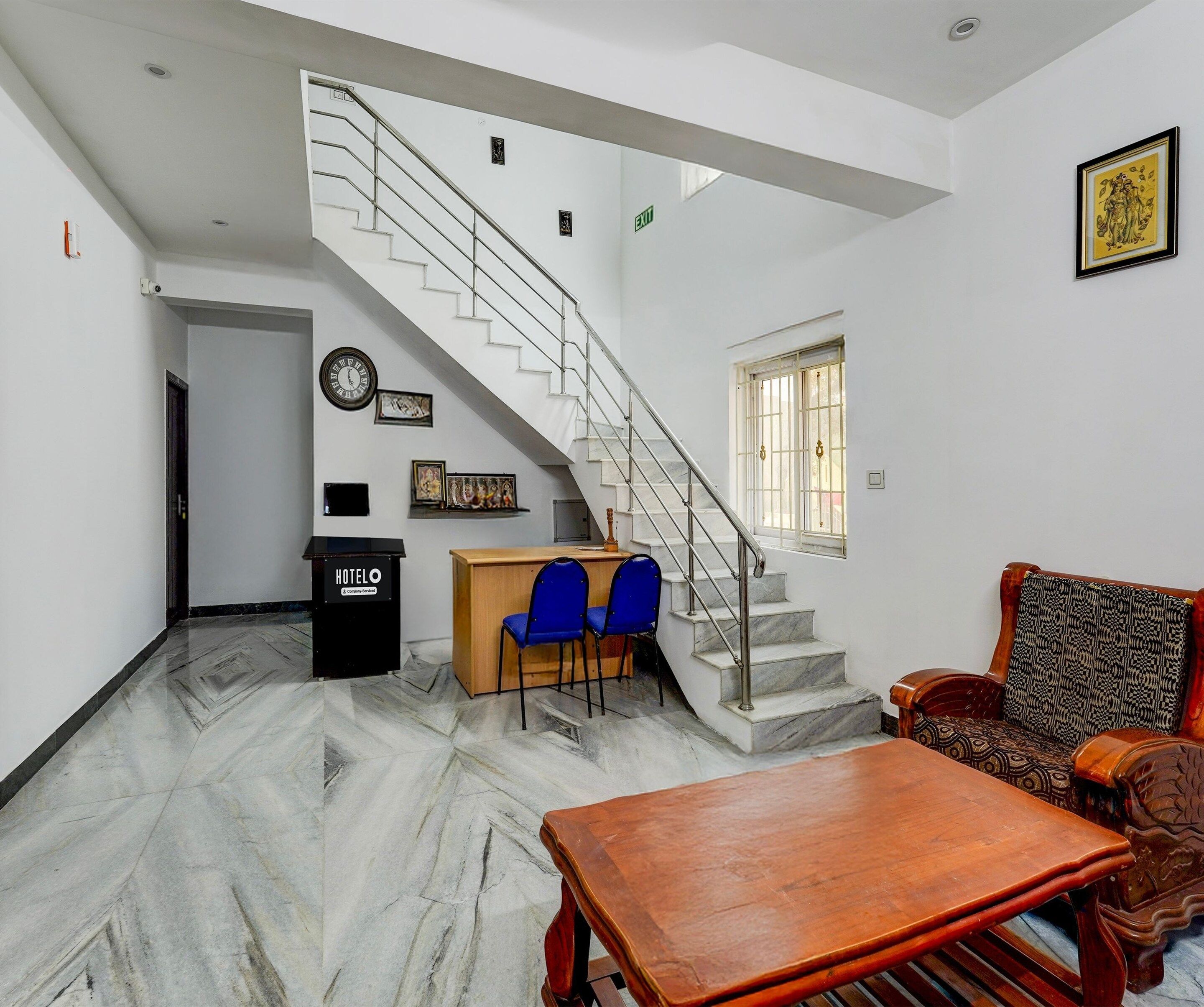 A hotel lobby with a marble floor, a wooden reception desk, blue chairs, a wooden sofa, a staircase, and a framed picture on the wall.