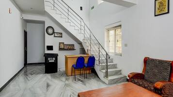A hotel lobby with a marble floor, a wooden reception desk, blue chairs, a wooden sofa, a staircase, and a framed picture on the wall.