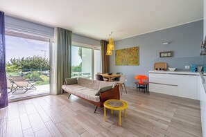 A modern living room with a sofa, dining table, and chairs, a kitchen area with cabinets and a countertop, and a balcony with a view of greenery. - VerdeBlù - Home Design by the Sea (Arma di Taggia)