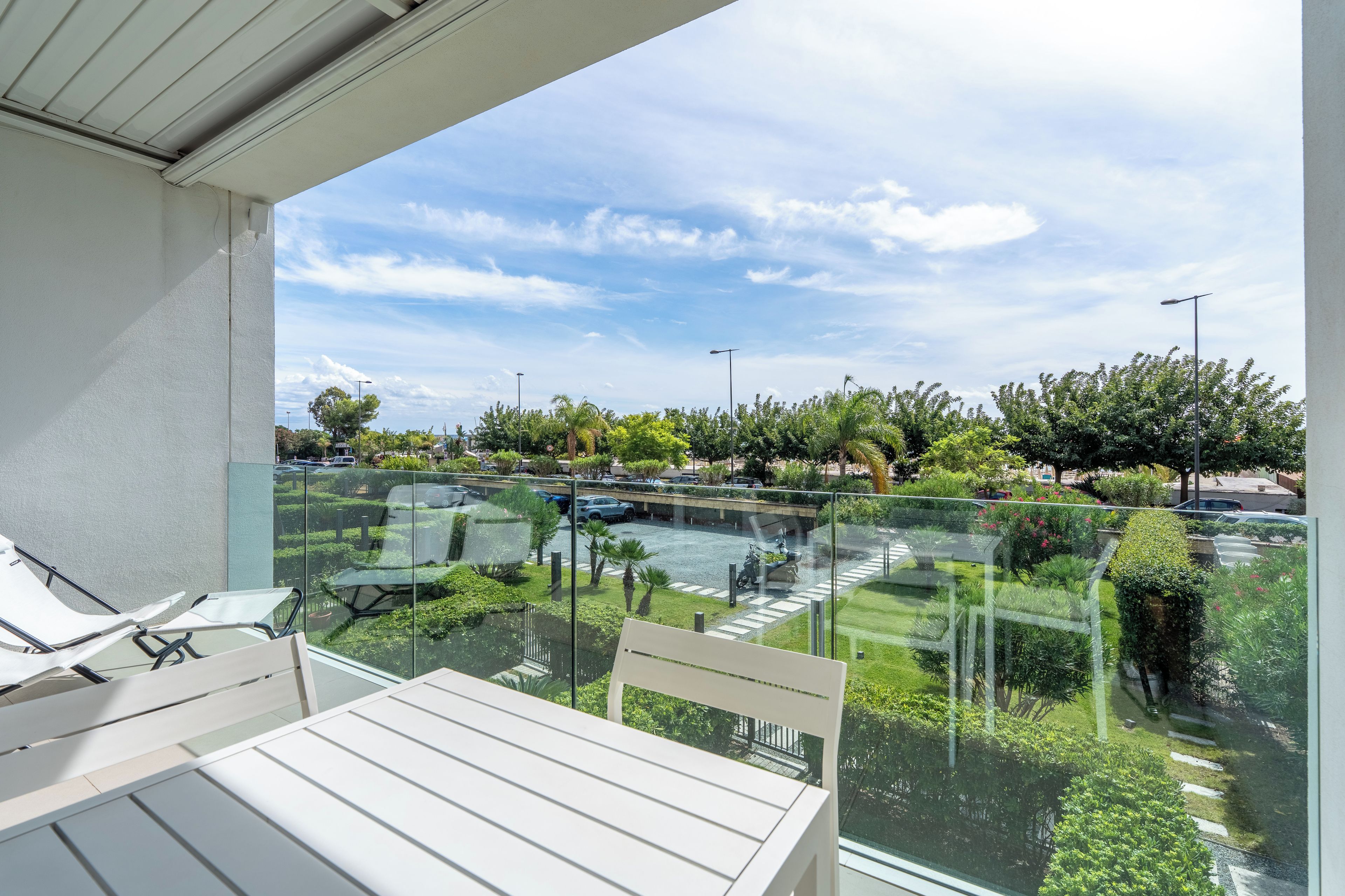 A balcony with a glass railing overlooking a landscaped area with trees and a parking lot.