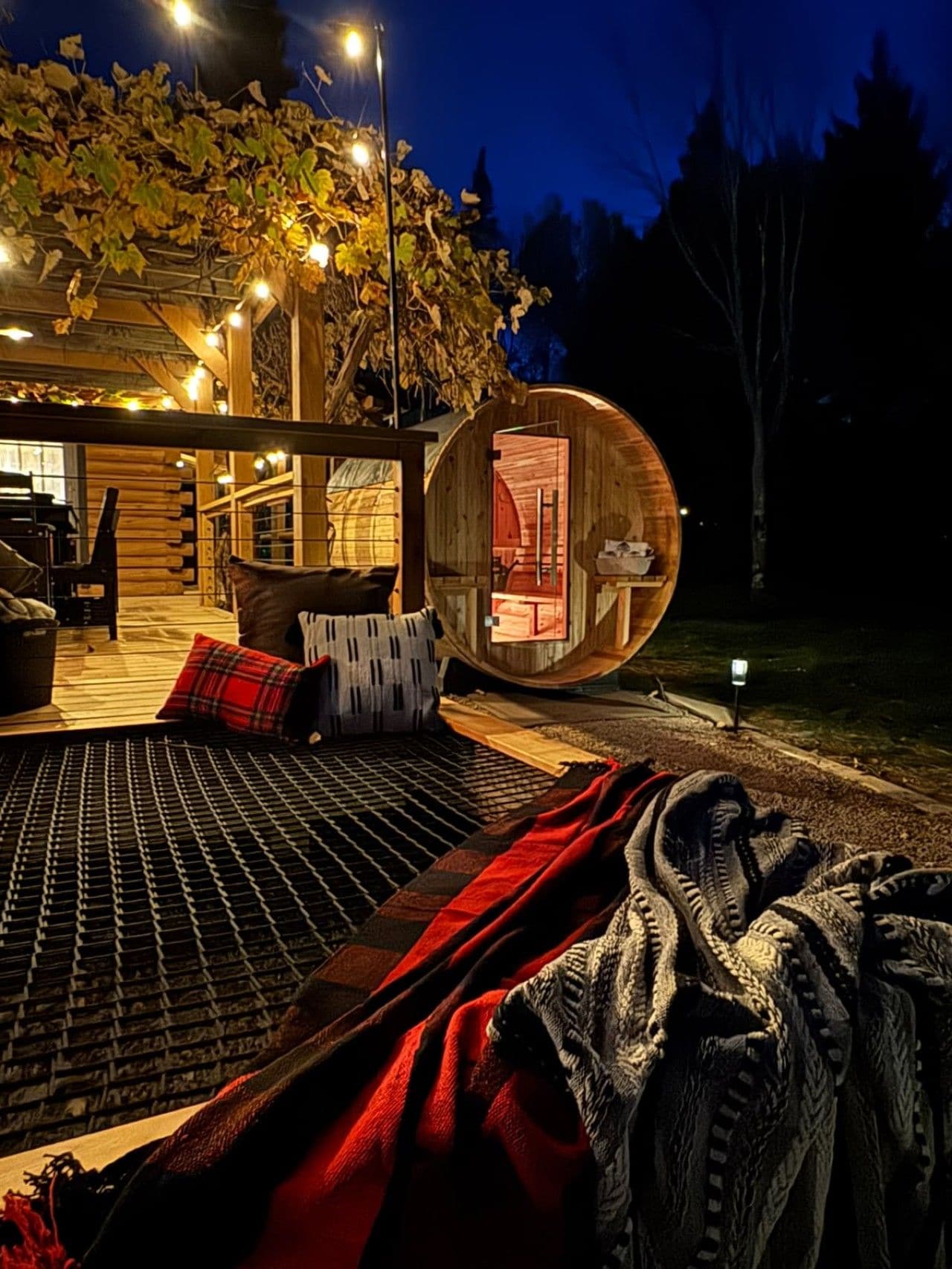 Un espacio de estar al aire libre acogedor, con un banco de madera, una manta estampada en rojo y blanco, y una estructura de barril de madera.