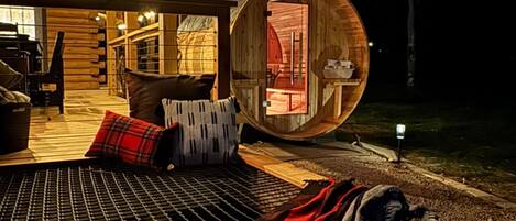 Un espacio de estar al aire libre acogedor, con un banco de madera, una manta estampada en rojo y blanco, y una estructura de barril de madera.