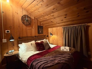 A wooden-paneled room with a bed, a wall clock, a lamp, and a curtain. - TheHiddenPond|Sauna|Hottub| Stargazing|Fishing|Ski (Windham)