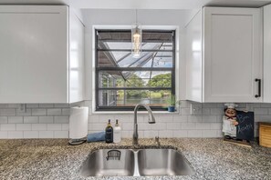 A kitchen with a double sink, a window with a view of greenery, and a chef figurine holding a sign. - Private Pool + Bar - Pet Friendly - Sleeps 6 (Port Orange)