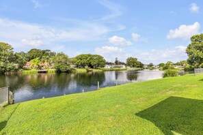 A serene lakeside view with a grassy foreground, a calm water body, and residential buildings in the background. - Private Pool + Bar - Pet Friendly - Sleeps 6 (Port Orange)