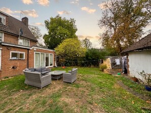 A residential backyard with a brick house, a patio with wicker furniture, and a garden area. - Soothing bedroom house in charming London neighborhood (London)