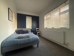Een slaapkamer met een bed, een stoel, een radiator en twee ramen met jaloezieën.