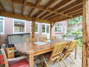 A covered outdoor dining area with a wooden table and chairs, a stone floor, and a view of a pink building with windows. - The Studio @ 18 (Kinross)