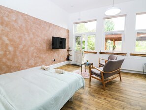 A bedroom with a bed, a chair, a TV, and large windows. - The Studio @ 18 (Kinross)