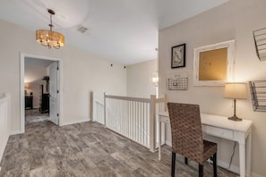 A hallway with a white railing, a desk with a lamp, and a wicker chair. - Luxury NSB Beach House | Private Pool & Bar (New Smyrna Beach)