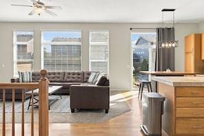 Ein modernes Wohnzimmer mit einer Couch, Couchtisch und einem Essbereich mit Holztisch und Stühlen. Es gibt eine Küchenzeile mit einem Edelstahlkühlschrank und Holzschränken. Große Fenster bieten einen Blick nach außen.
