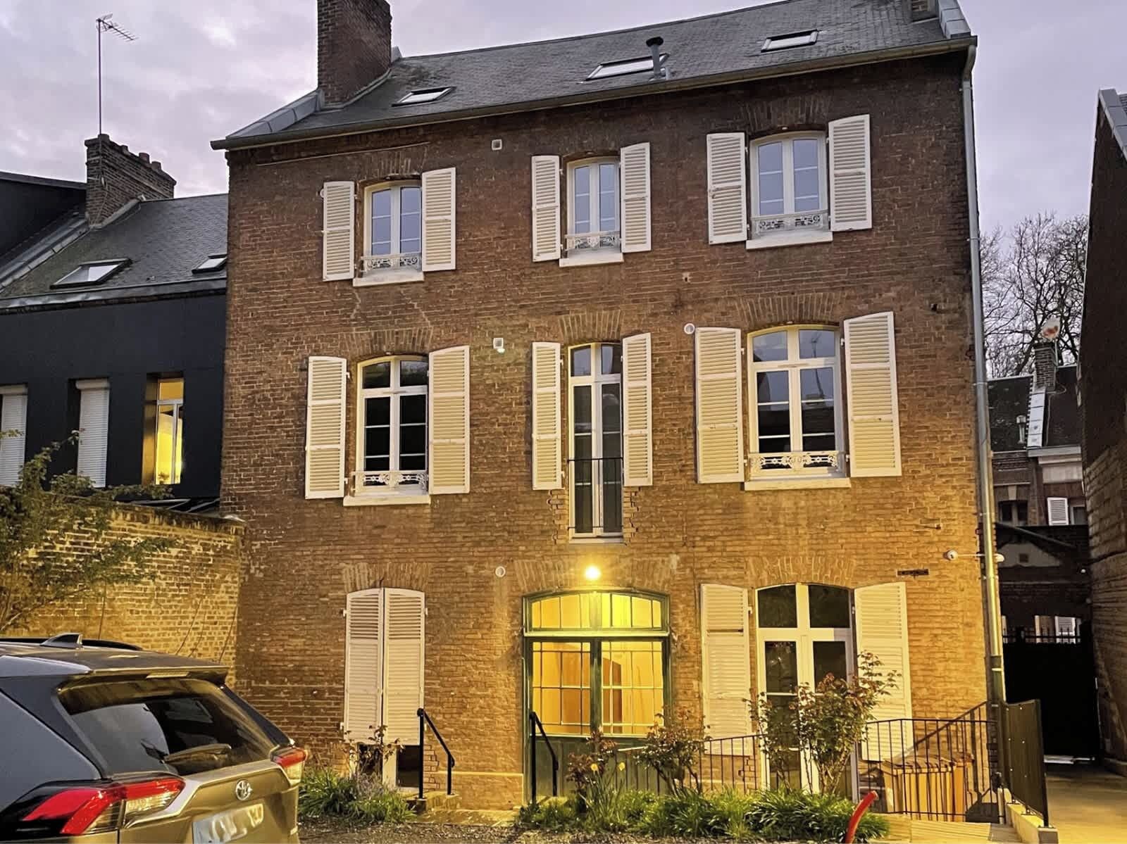 Un immeuble en briques de plusieurs étages, doté de volets blancs, une entrée centrale avec une porte en verre et une voiture garée devant.