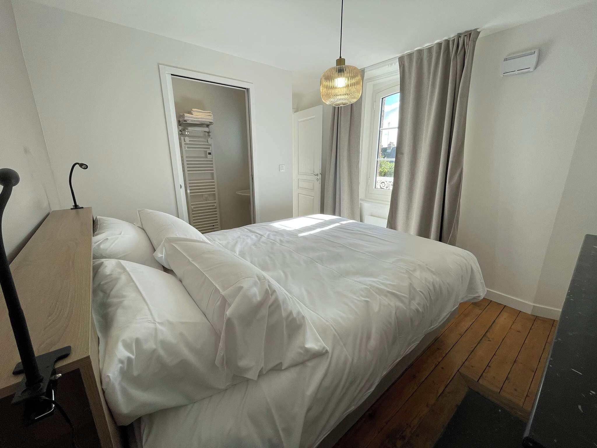 Une chambre à coucher comprenant un lit, une table de chevet, un luminaire suspendu, une armoire et une fenêtre avec des rideaux.
