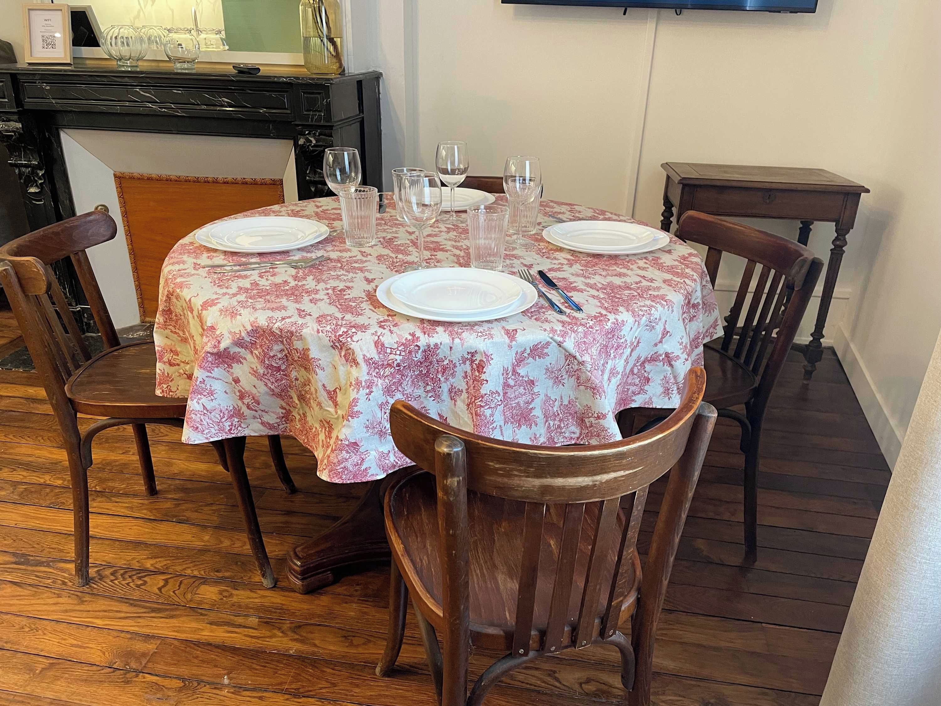 Une table à manger dressée pour quatre personnes, avec une nappe à motifs floraux, des chaises en bois et une cheminée en arrière-plan.