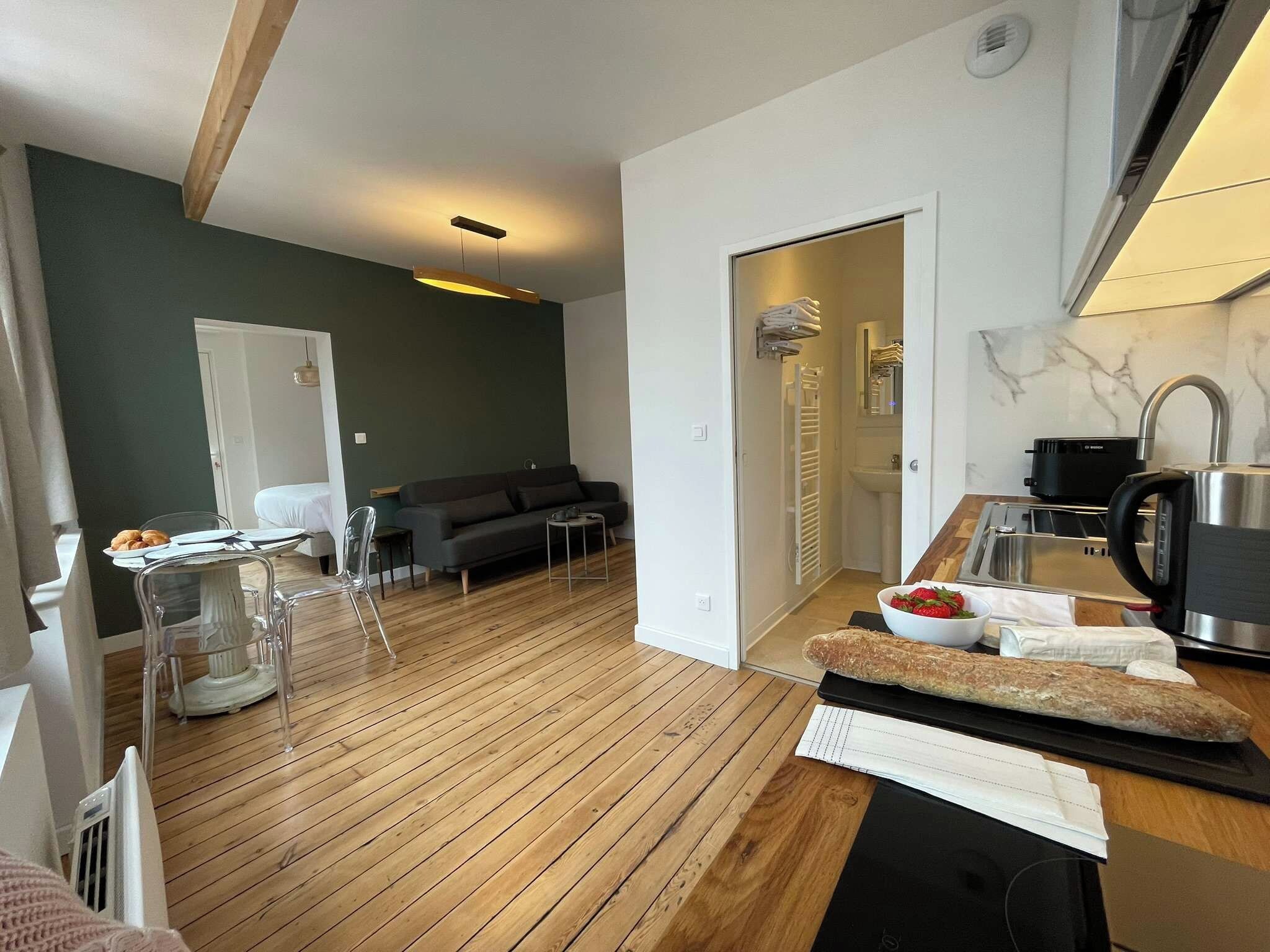 A modern living room with a wooden floor, a sofa, a dining table with chairs, and a kitchen area with a kettle and a bowl of strawberries.