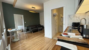 A modern living room with a wooden floor, a sofa, a dining table with chairs, and a kitchen area with a kettle and a bowl of strawberries.