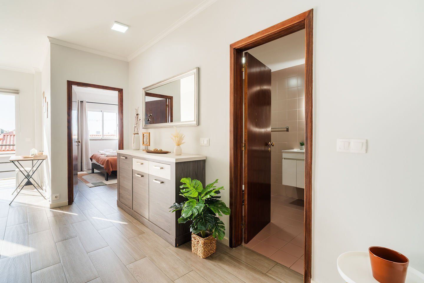 A hotel room with a bed, a small desk, a mirror, a potted plant, and a bathroom with a towel rack.