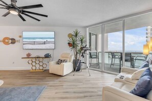 A modern living room with a large window, a ceiling fan, a mounted TV, and a seating area with a view of the ocean. - 20th Floor Luxury Condo | Halifax River Views (Holly Hill)