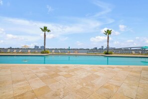 Piscina com espreguiçadeiras e palmeiras, com um horizonte urbano e uma ponte ao fundo.