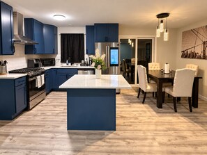 A modern kitchen with blue cabinets, a central island, stainless steel appliances, and a dining area with a chandelier. - Spacious 4BR Retreat - Game Room, Full Kitchen, Private workspace & Four Seasons (Rockford)