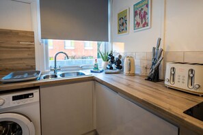 A modern kitchen with a built-in washing machine, a toaster, and a window with a view of a brick building. - 3-bed flat by Felixstowe seafront with roof terrace and parking (Felixstowe)