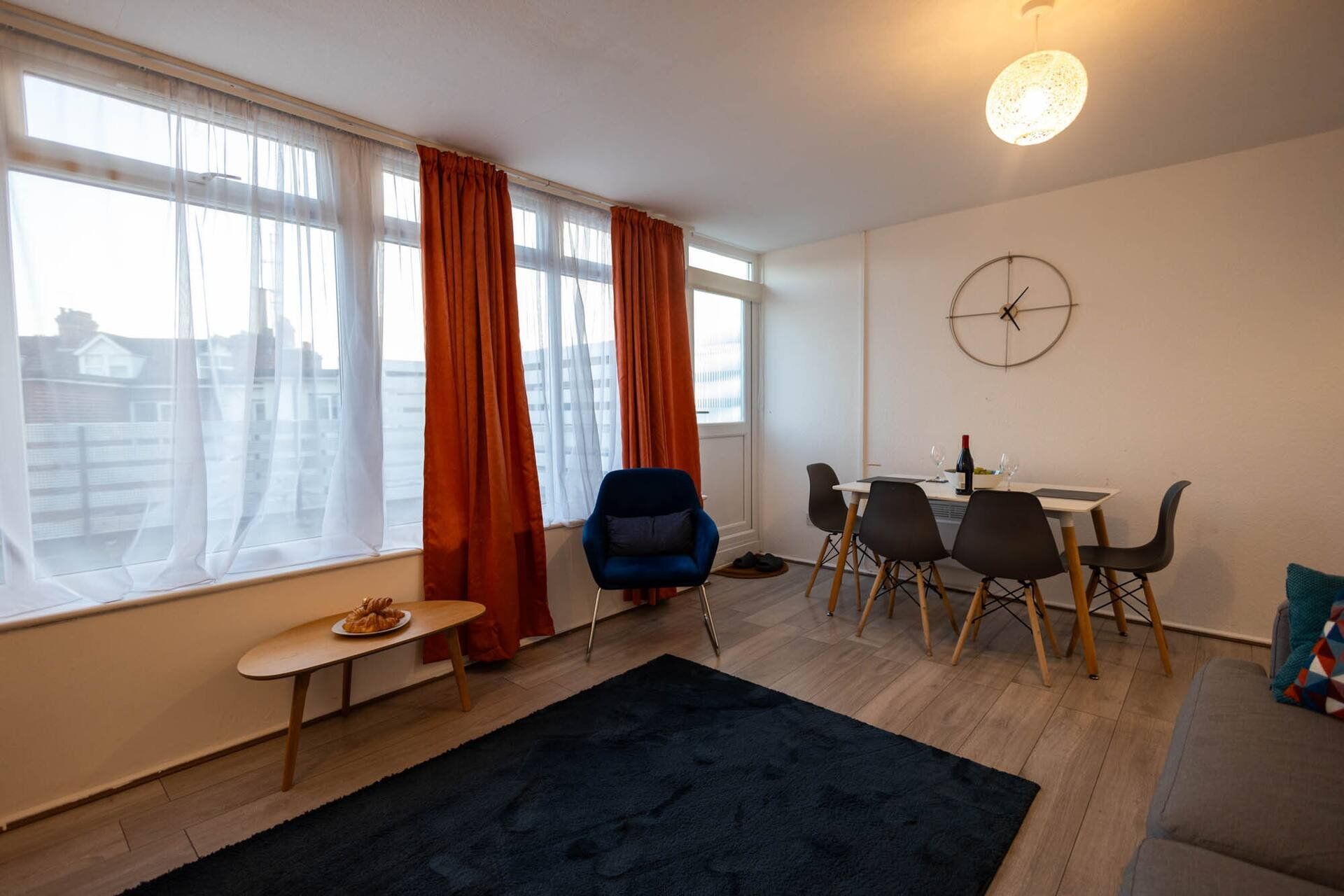 A modern living room with a dining area, large windows with curtains, a round wall clock, and a small table with a plate of food.