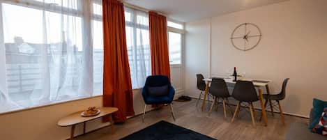 A modern living room with a dining area, large windows with curtains, a round wall clock, and a small table with a plate of food.