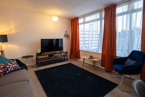A modern living room with a flat-screen TV, a sofa, a blue armchair, a small table, and a large window with curtains. - 3-bed flat by Felixstowe seafront with roof terrace and parking (Felixstowe)