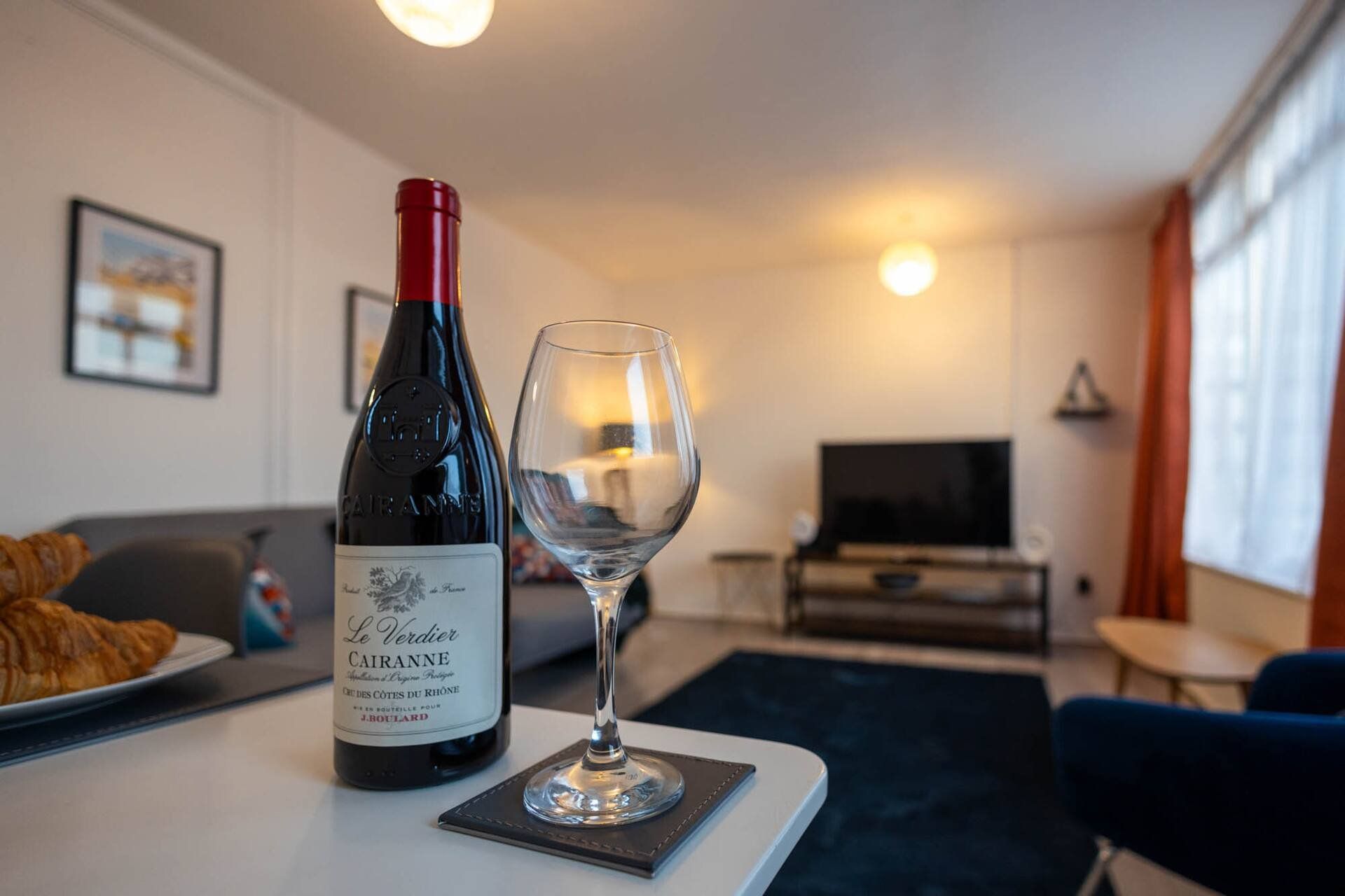 Eine Weinflasche und ein Glas auf einem Tisch in einem Wohnzimmer mit Sofa, Fernseher und einem fenster mit Vorhängen.