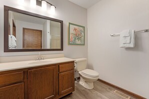 Une salle de bain avec un meuble-lavabo en bois, des toilettes blanches, un grand miroir avec éclairage et un tableau floral encadré accroché au mur.