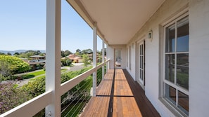 Un balcón techado con cubierta de madera, barandales blancos y amplios ventanales que ofrecen vistas a una zona residencial y colinas.