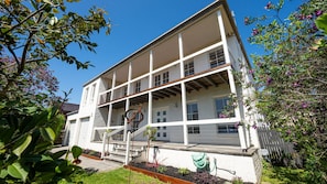 Ein zweistöckiges Gebäude mit Balkon und einem Gartenbereich mit Pflanzen, vor einem strahlend blauen Himmel.