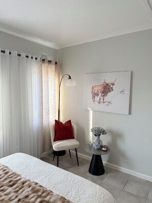 Quarto com cama, cadeira, luminária, um vaso com flores e um quadro na parede.