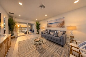 A modern living room with a grey sofa, a glass coffee table, and a painting of a beach scene. - 2-bedroom 2- bath Waterfront Paradise With Intracoastal Views & Dock (West Palm Beach)