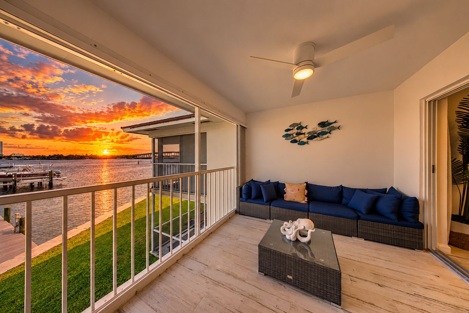 A balcony with a view of a sunset over a body of water, a wicker sofa with cushions, and a small table with a decorative item.