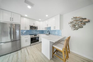Een moderne keuken met witte kastjes, roestvrijstalen apparatuur, een blauwe spatwand en een houten stoel.