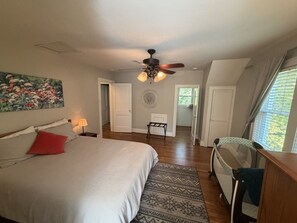 Une chambre à coucher avec un lit, un ventilateur de plafond, un tableau, une table de chevet avec une lampe et une vue sur un couloir.