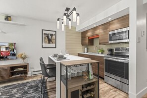 Ett modernt kök med en central köksö, rostfria vitvaror och en matplats med en svart stol och ett litet bord.