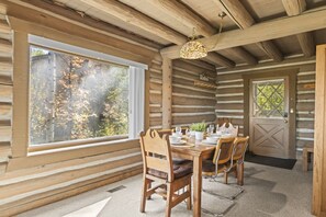 Ein Interieur einer Blockhütte mit einem Esstisch, gedeckt für vier Personen, Holzstühlen, einer Chandelierleuchte und einem großen Fenster mit Blick auf Bäume.