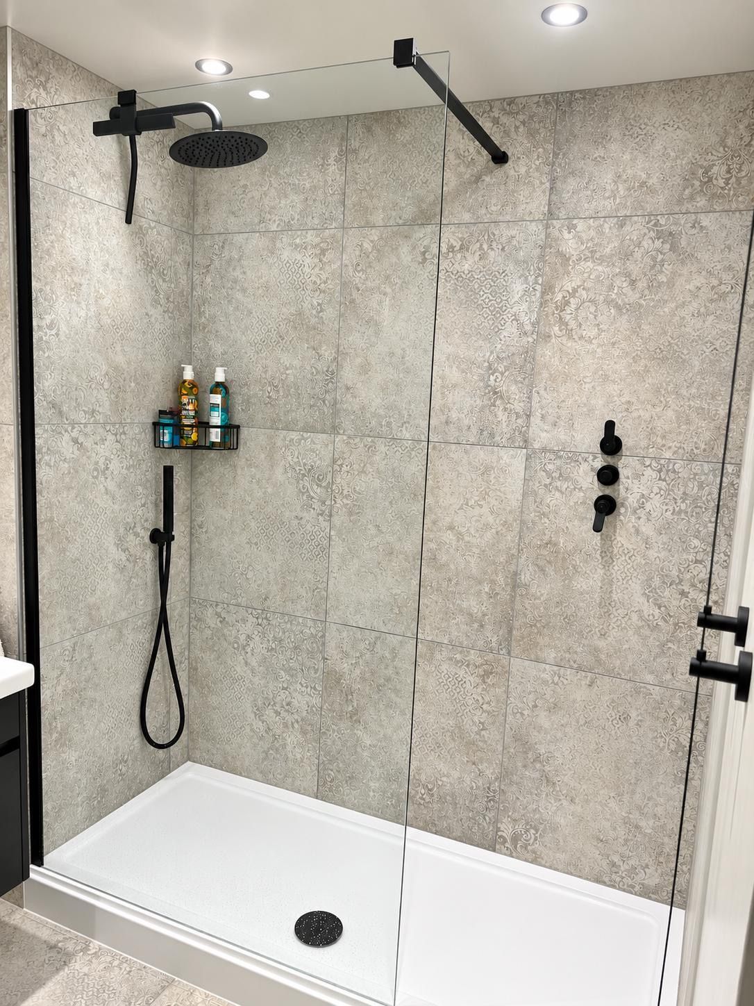 A walk-in shower with a glass door, a handheld showerhead, a wall-mounted control panel, and a shelf with toiletries.